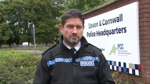 BBC Acting Chief Constable Jim Colwell pictured. He is stood in front of a white sign reading: "Devon and Cornwall Police Headquarters." He has short dark hair that has been swept back. He has a short beard. He is wearing a black police jacket with blue and white check pattering. There is also a blue badge on his chest that reads "police".