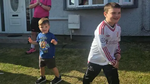 Dáithí is playing in a front garden with another young boy. Has short brown hair, wearing a white long sleeve Liverpool jersey and black tracksutt bottoms. The other young boy has short brown hair, wearing a blue t-shirt, dark shorts and trainers. A man is in the background, he is wearing a pink GAA jersey, black shorts and pink trainers. He is holding his hands together.
