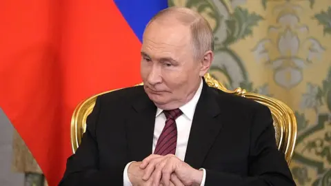 PAVEL BEDNYAKOV/POOL/AFP via Getty Vladimir Putin sitting in a gold chair, hands clasped and looking down. The Russian flag is draped behind him.