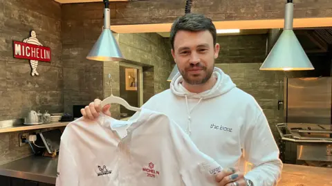 Liam Dillon is wearing a white hoody with the words, the boat written on the left side of his chest. He is holding a chef's uniform on a hanger and standing in a restaurant. On the wall there is the Michelin logo