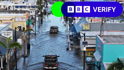 Getty Images Cars in Florida drive through a street flooded with water after Hurricane Helene.