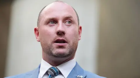 PA Media A bald man, wearing a blue suit and blue stripy tie, looks to his left in a close-up shot 