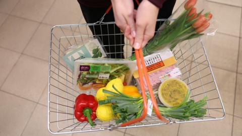 A shopping basket full of fruit and vegetables is held by a supermarket customer.