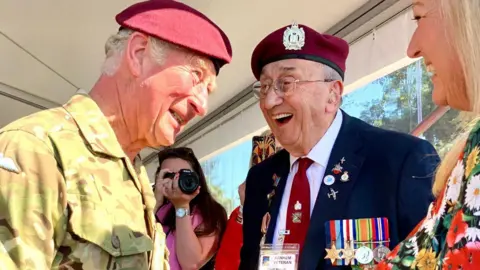 King Charles and Geoffrey Roberts are speaking with each other, both are smiling and laughing. Mr Roberts is wearing a suit and his medal ribbon.