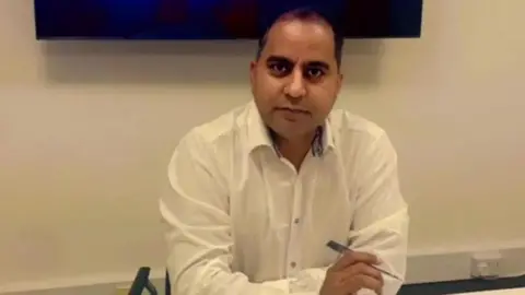 Arshad Mahmood Arshad Mahmood, who has black hair and is wearing a white shirt, sits at a table holding a pen. He is sat in front of a white background.
