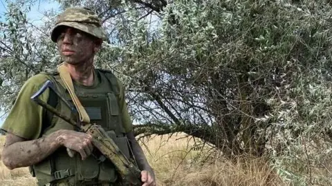 Maksym is standing outside holding a rifle, he has camouflage paint on his face and arms. He is wearing a green t-shirt and combat vest, holding a rifle and looking to his right.
