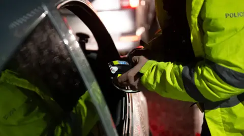 A police officer wearing hi-viz holds a breathalyser device through the driver's window of a car.