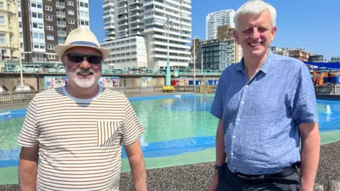 Two men are standing in front of a paddling pool, with blocks of flats in the background. The man on the left has a beard and sunglasses on, and wearing a striped white and brown t-shirt and a white hat. The man on the right has a blue short sleeved shirt and white hair
