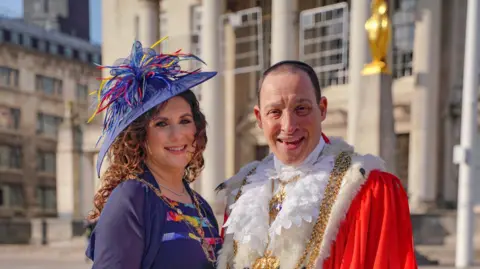 Leeds City Council Picture shows Elayna Cohen on the left wearing a blue suit, with a small golden chain round her neck and a blue hat.
Cllr Cohen is pictured to the right wearing the Lord Mayor dress robes, red with a white collar and a golden chain.
Both are stood infront of the Civic Hall in Leeds.