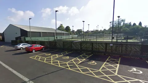 A number of tennis courts beyond a fence, with several floodlights. On the near side of the fence is a car park with two cars. A number of spaces are designated disabled bays.
