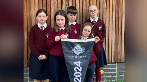 five children wearing burgundy school uniforms are standing together, they each have sad expressions on their face. Two girls at the front are holding a flag which says 'Tá Cúpla Focal Scoil Spreagtha 2024'. Behind them is a wall with some wooden panelling.  