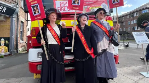 Three women (Sam Strudwick, Lillian Francis and Carol King) are chained to the front of the fire engine. They wear long Edwardian women's coats and hats in the suffragette style. They are holding up placards.