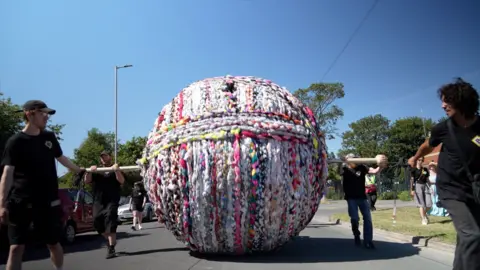 A giant ball of yarn is rolled along a road by people holding a piece of wood which runs through the ball.