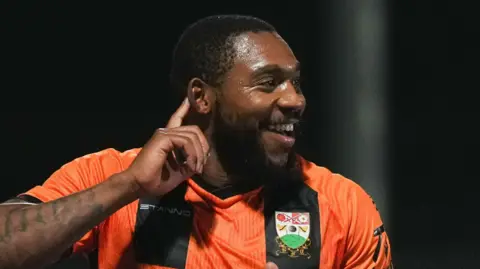 Britt Assombalonga in an amber and black Barnet FC jersey celebrates a goal during a match.