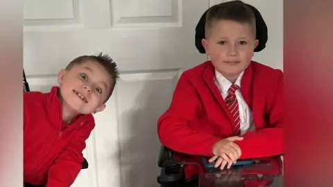 WellChild Two boys wearing red school uniforms smile at the camera while sitting side by side in front of a white panelled door. The boy on the right is seated in an electric wheelchair with a joystick control visible, wearing a shirt and striped tie under his red cardigan. The boy on the left leans in towards his brother, wearing a red zip-up fleece.
