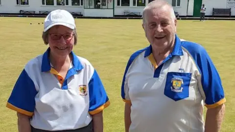 Troy Mustow Two people standing next to each other in blue and white bowling polo shirts on a green lawn. They are smiling widely in sunshine.