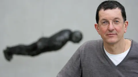 PA Media Sir Antony Gormley, who has short black hair and rounded spectacles, poses for a picture with a sculpture based upon his own body hanging in the background.