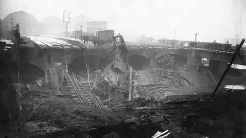 Leeds Museums A black and white image showing a railway bridge where a large section of the structure has collapsed into a deep cavity below. The image has a heavy, smoky appearance, consistent with the aftermath of a fire.