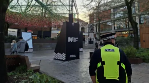 Norfolk Police Hay Hill in Norwich. The space is seated with planting including trees. people are walking through including a police officer wearing a high-vis vest. Shops surround the space.