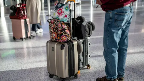 Two people are standing in an airport holding suitcases with bags sitting in top. 