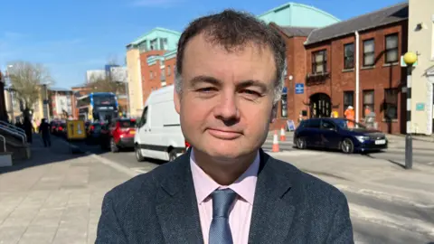 Councillor Gary Ridley wears a grey suit, pink shirt and grey tie. He smiles at the camera.