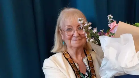 Councillor Eacock is wearing a white jacket with a large gold chain around her neck. She is being handed flowers to mark the occasion.
