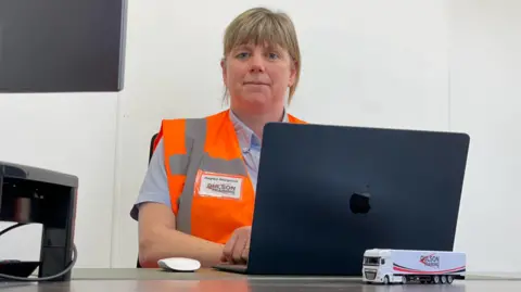 A woman wearing an orange high-vis vest sat behind a laptop with plain walls behind her.