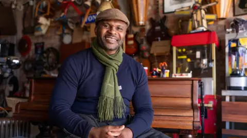 A man in a blue jumper, green scarf and brown flat cap smiles at the camera. There is a piano and cabinets with toys and trinkets in behind him. 
