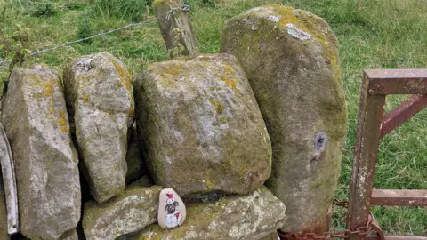 Stephanie Marie Winter A small painted rock depicting a cartoon sheep is placed on a stonewall in a field.