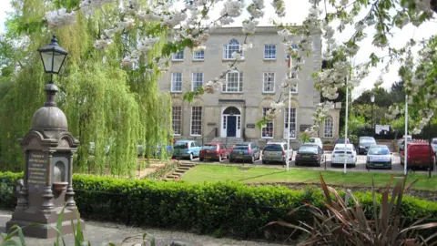 A large three storey Georgian-style house, in a cream colour with about 12 vehicles in front of it. The photographer has taken the photo whilst looking through a blossom tree in the foreground, with other mature trees nearby and a lawn in front of the building. 