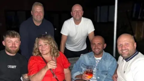 Family A group photo shows a woman in a red dress surrounded by five men. Three of the men are sitting on either side of her while two are standing beside her. They are all smiling.