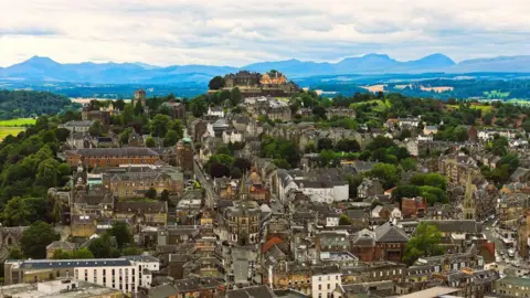 Kevin Hamill Stirling Castle towering over houses on its hilltop perch, under a cloudy sky, with hills in the distance.