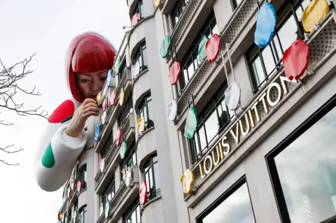 Reuters Exterior of Louis Vuitton store with logo and a giant artificial figure of Yayoi Kusama leaning over the roof and apparently attaching multi-coloured polka dots to the building