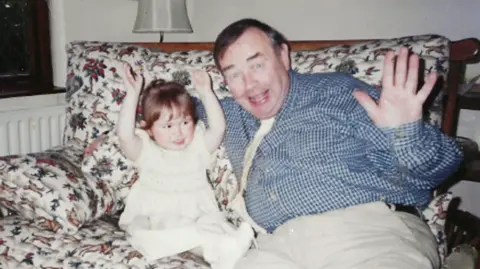 McGrath family Sean McGrath, 61, with his two-year-old granddaughter Sara.  They are sitting on a sofa smiling and waving at the camera.   Sean has short, dark hair and is wearing a blue checked shirt and beige trousers.   Sara has dark hair and is wearing a white dress. 