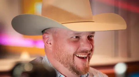 Cody Johnson is smiling with a cream cowboy hat on. He has a ginger beard and brown eyes with studio lighting behind him. 