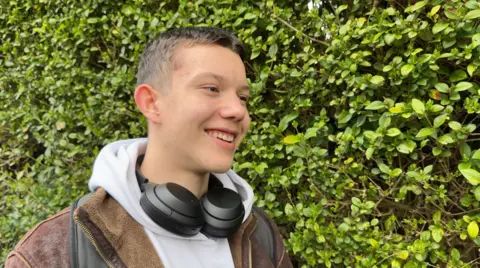 Image shows a student called Mavi. He is looking off to the left side. He has brown short hair. He is smiling. He had a white hoodie on and a brown jacket over the top. You can see the straps of his backpack which are black. He has a pair of black headphones round his neck. The whole background is filled with a large green bush. 