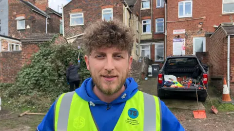 A close-up of a man with curly brown hair looking at the camera. He wears a blue hoodie and yellow hi-viz jacket with a blue TS logo on it.