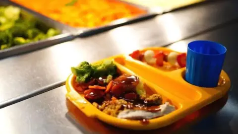 A yellow school tray of food with separate sections filled with chicken, broccoli and gravy and a blue beaker. 