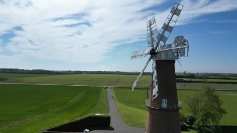 BBC/Joe Bilton A six-sailed windmill photographed from the skies. It has white sails and a red-brick body. It is surrounded by green fields.