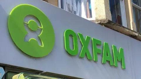 The sign above an Oxfam shop. The name and logo are cut from green plastic on a white background.