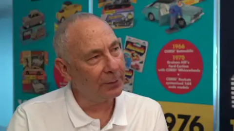 A man in a white t-shirt with grey hair looks at the camera