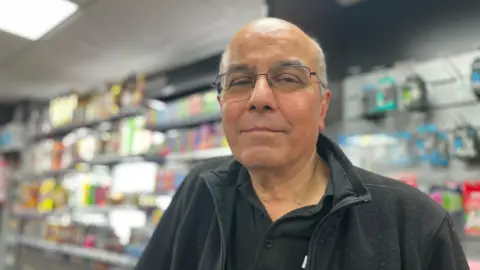 Emma Baugh/BBC Vidyut Soni looks directly at the camera, unsmiling. He is wearing glasses and a black polo shirt and fleece. Behind him are shelves stocked with goods. 