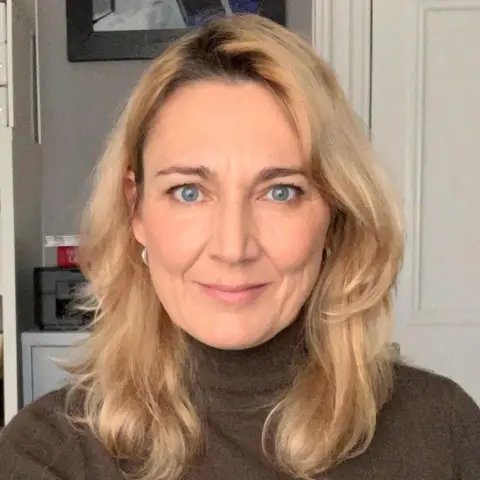 Leasha Esslemont Leasha Esslemont, a woman with shoulder-length, blonde hair wearing a brown turtleneck top. The white door and a painting are visible on the background. 