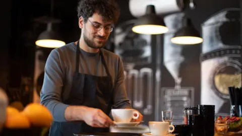 Getty Images Barista with curly hair wearing glasses hands a coffee to a customer in a white cup with a saucer