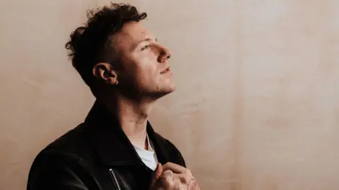 Callum Beattie has dark hair and is pictured in profile wearing a dark jacket over a white T-shirt. His head is titled upwards as he looks off to the distance.