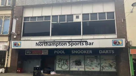 A view of the entrance to the Northampton Sports Bar. The windows of the former bar are covered in graffiti and there is some rubbish outside the door. Windows above the entrance have been blacked out. 