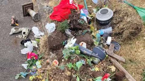 Louise Hirst The contents of a bin, including broken ornaments, vases, graveside flower holders, grass, plants and soil, are strewn about by the side of a gravel path.