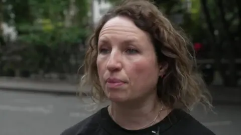 A woman with curly hair wearing a black top and microphone headset is speaking on a city street, with trees and railings in the background.