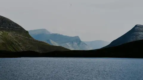 Milo Beswick A landscape featuring a calm body of water in the foreground and a range of mountains in the background under a muted sky. 