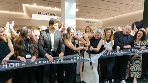 Sephora A group of about 20 people stand behind a giant ribbon while a woman cuts it with a giant pair of scissors.
